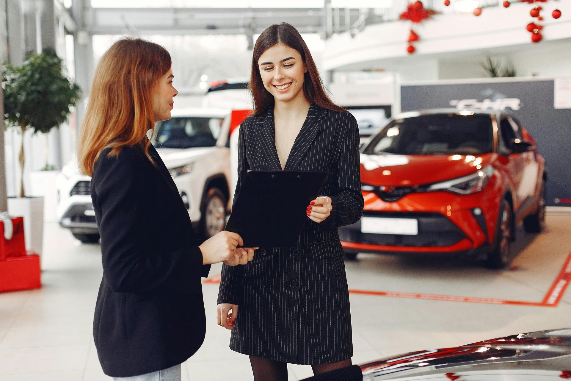 Ein fröhlicher, junger und freundlicher Autohändler in elegantem, schwarzem Kleid zeigt einer lächelnden Kundin in schwarzer Jacke einen Vertrag, während er in einem Autohaus vor neuen, glänzenden Fahrzeugen steht.