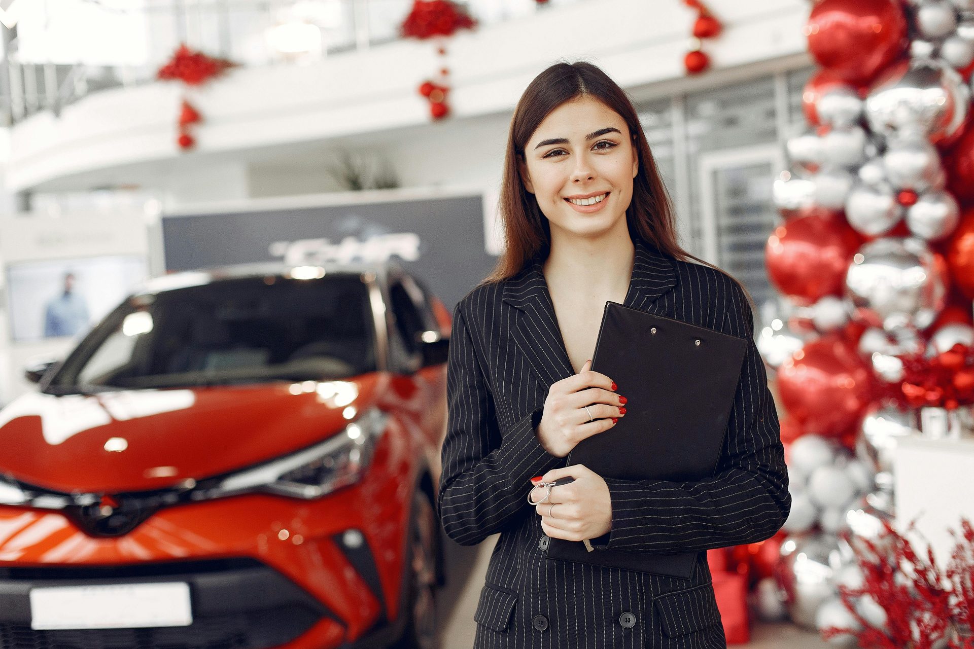 Eine fröhliche, freundliche und stilvolle Verkäuferin in einem eleganten schwarzen Abendkleid steht mit Klemmbrett in einem Autohaus vor einem neuen, glänzend roten Wagen und blickt in die Kamera.